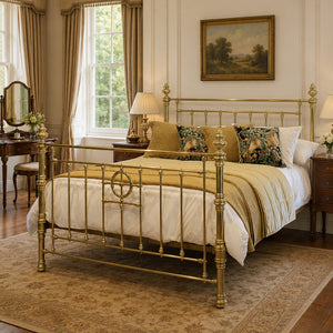 A classic brass antique bed frame with ornate turned posts and a decorative centre panel, styled in a traditional bedroom with white bedding, gold-toned pillows, and a patterned rug.