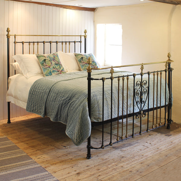 An antique metal bed frame with ornate black scrollwork and brass/gold accents, dressed in white bedding and a textured sage green quilt, set in a rustic bedroom with a wooden floor and white panelled wall.