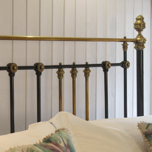 Close-up of an ornate metal bed frame headboard with black and brass vertical bars, decorative finials, and a horizontal brass rail, shown against a white paneled wall with white bedding and patterned pillows.