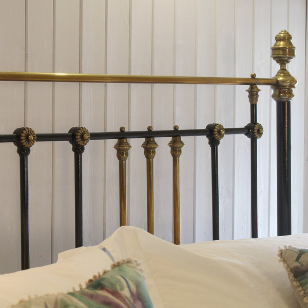 Close-up of an ornate metal bed frame headboard with black and brass vertical bars, decorative finials, and a horizontal brass rail, shown against a white paneled wall with white bedding and patterned pillows.