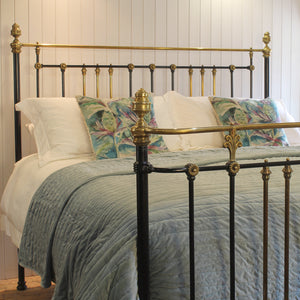 A bed with a black metal frame featuring ornate gold brass accents on the headboard and footboard, dressed in white linens, a light blue-gray quilt, and two tropical patterned accent pillows against a white paneled wall.