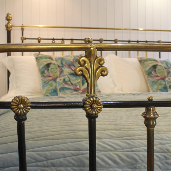 Close-up of an ornate metal bed frame with gold-toned brass accents and black bars, dressed in a light blue quilted bedspread and patterned pillows against a white paneled wall.