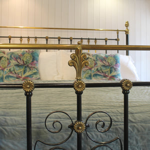 Close-up of an ornate metal bed frame with a black wrought-iron style base and gold-toned accents, including a matching headboard. White pillows and floral-patterned bedding are visible against a white paneled wall.