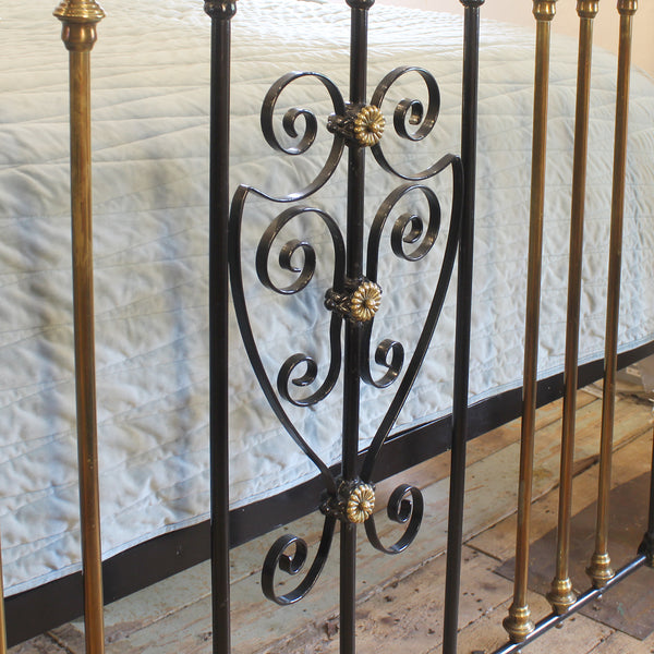 Close-up of an ornate metal bed frame with black scrollwork and gold-toned decorative accents, shown beside gold-toned posts on a bed with a light quilted cover.