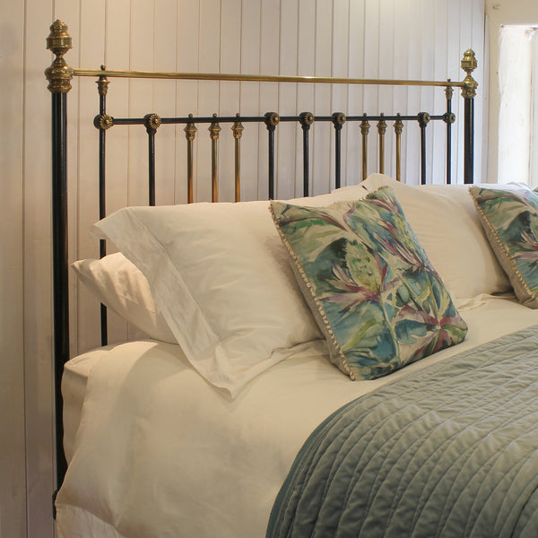 A brass and black metal bed frame with decorative finials, styled with white bedding, patterned tropical accent pillows, and a textured teal quilt in a bedroom setting.