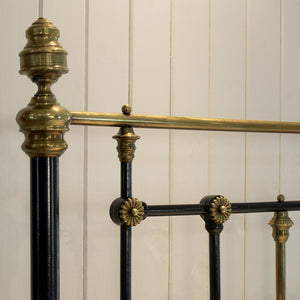 Close-up of an ornate metal bed frame with black painted vertical posts and polished brass or gold-toned horizontal bars and decorative finials, set against a white paneled wall.