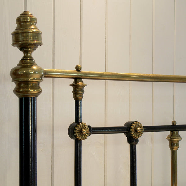 Close-up of an ornate metal bed frame with black painted vertical posts and polished brass or gold-toned horizontal bars and decorative finials, set against a white paneled wall.