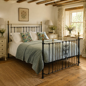A black wrought-iron antique metal bed frame with brass accents, styled in a rustic bedroom with a green quilt, white bedding, and patterned pillows.