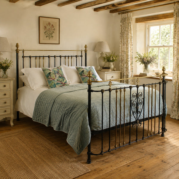 A black wrought-iron antique metal bed frame with brass accents, styled in a rustic bedroom with a green quilt, white bedding, and patterned pillows.