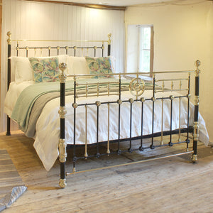 An ornate metal bed frame with black and gold-toned vertical posts and decorative scrollwork, shown made up in a rustic bedroom with white bedding, green throw blanket, and two tropical print pillows.