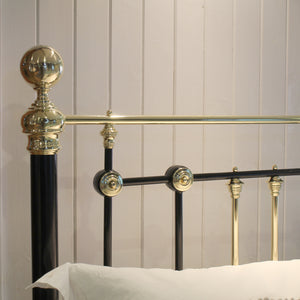 Close-up of a metal bed frame with a black vertical post and ornate gold-toned accents, including a spherical finial, decorative round connectors, and a horizontal rail, set against a light paneled wall with a white pillow visible at the bottom.