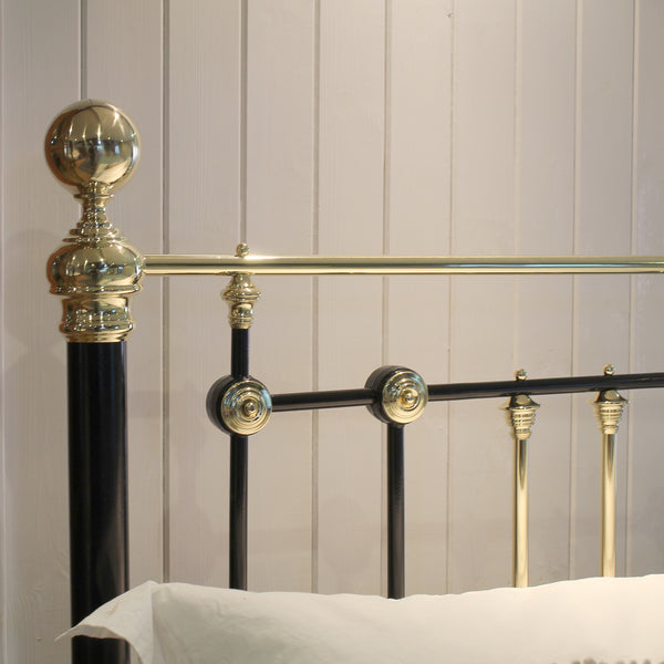 Close-up of a metal bed frame with a black vertical post and ornate gold-toned accents, including a spherical finial, decorative round connectors, and a horizontal rail, set against a light paneled wall with a white pillow visible at the bottom.