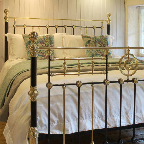 A classic black and gold metal bed frame with ornate gold accents, dressed in white bedding and green patterned pillows, set against a white paneled wall.