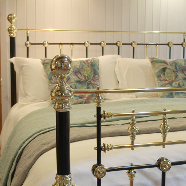 A close-up of a bed with a black metal frame featuring ornate gold-toned accents and a matching headboard, dressed in white bedding with a sage green throw and floral accent pillows.
