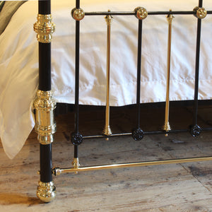 Close-up of a black and gold metal bed frame with ornate brass-like fittings, shown beside a bed with white bedding on a wooden floor.