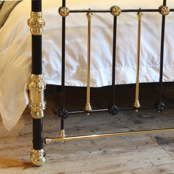 Close-up of a black and gold metal bed frame with ornate brass-like fittings, shown beside a bed with white bedding on a wooden floor.
