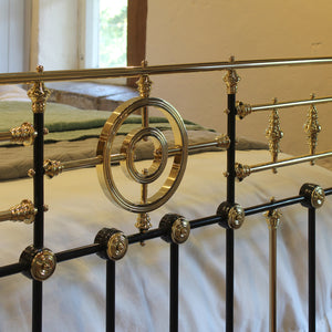 Close-up of an ornate metal bed frame with black vertical bars and polished gold-toned decorative rails and knobs, shown in a bedroom setting with bedding and a window in the background.