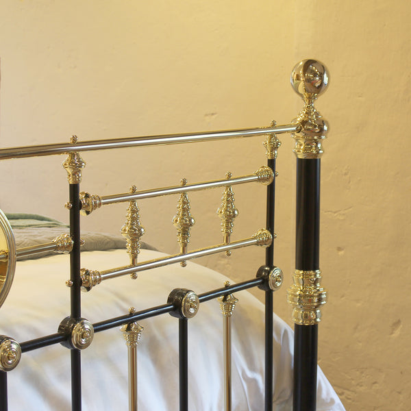 Close-up of a metal bed frame with black painted rails and ornate gold-toned decorative accents, shown against a warm beige wall with white bedding visible.