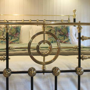 Close-up of an ornate metal bed frame with a black and polished brass/gold structure, shown in a bedroom setting with patterned pillows and white bedding.