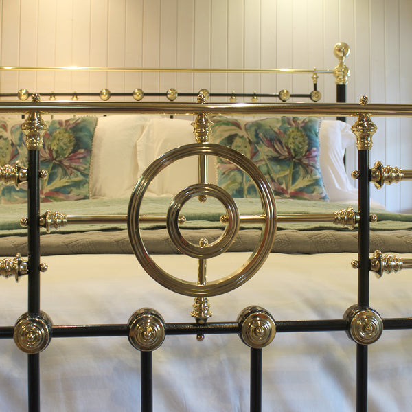 Close-up of an ornate metal bed frame with a black and polished brass/gold structure, shown in a bedroom setting with patterned pillows and white bedding.