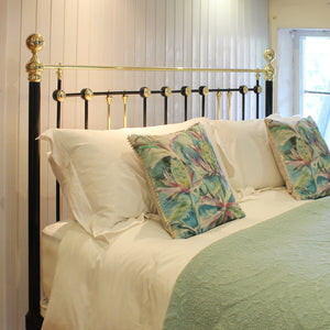 A bed dressed in white linens with two decorative tropical print pillows and a mint green throw, set against a black metal headboard with gold accents in a bedroom setting.