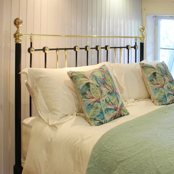 A bed dressed in white linens with two decorative tropical print pillows and a mint green throw, set against a black metal headboard with gold accents in a bedroom setting.