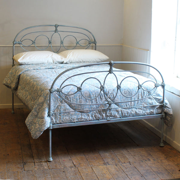 A vintage-style metal bed frame with an ornate circular headboard and footboard, dressed in a patterned blue-gray quilt and white pillows, set in a room with a rustic wooden floor and white walls.