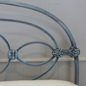 Close-up of an ornate metal headboard frame with a distressed blue-gray finish, showing curved rails and decorative scrollwork against a light wall.