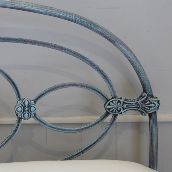 Close-up of an ornate metal headboard frame with a distressed blue-gray finish, showing curved rails and decorative scrollwork against a light wall.