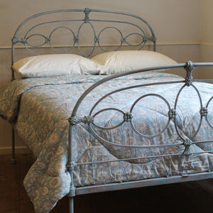 A metal bed frame with an ornate circular scroll design in a distressed blue-gray finish, shown with white pillows and a blue floral patterned quilt in a bedroom setting.