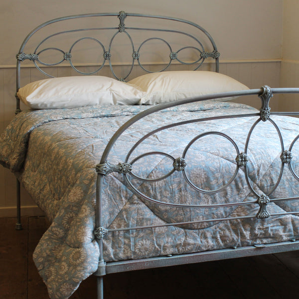 A metal bed frame with an ornate circular scroll design in a distressed blue-gray finish, shown with white pillows and a blue floral patterned quilt in a bedroom setting.