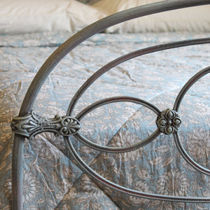 Close-up of a metal bed frame with decorative scrollwork and curved bars, shown on a bed with a blue floral quilt and white pillows in a bedroom setting.