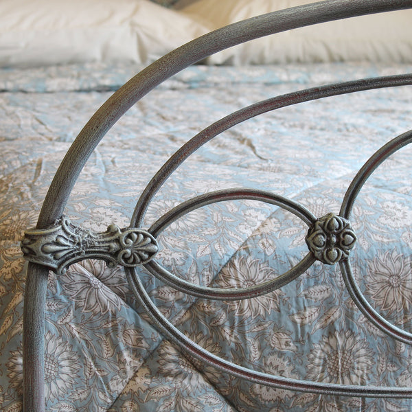 Close-up of a metal bed frame with decorative scrollwork and curved bars, shown on a bed with a blue floral quilt and white pillows in a bedroom setting.