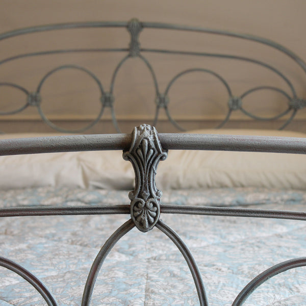 Close-up of a metal bed frame with an ornate decorative bracket and circular scrollwork, shown on a bed with patterned bedding in a softly lit bedroom.