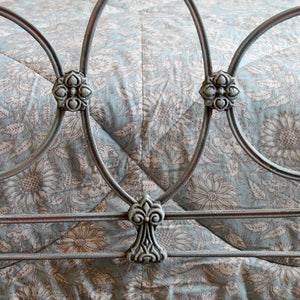 Close-up of a metal bed frame with circular and scroll decorative elements, shown against a light blue floral quilted bedding background.