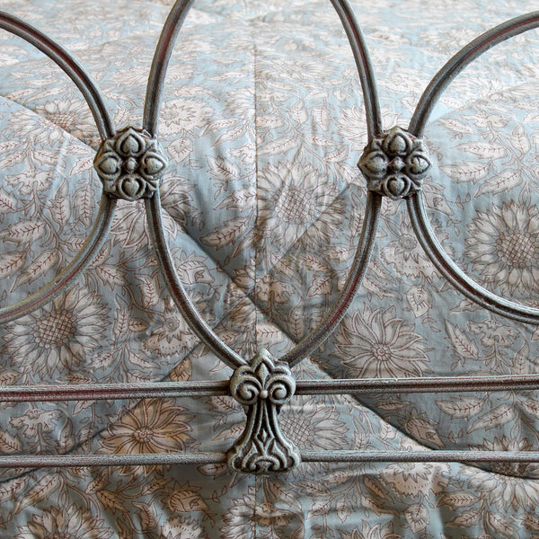Close-up of a metal bed frame with circular and scroll decorative elements, shown against a light blue floral quilted bedding background.