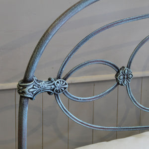 Close-up of an ornate metal bed frame headboard with curved scrollwork and decorative floral accents, shown against a light paneled wall.