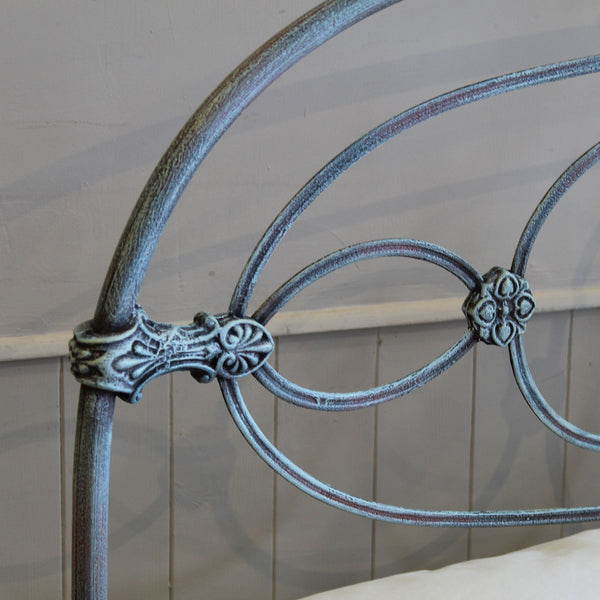 Close-up of an ornate metal bed frame headboard with curved scrollwork and decorative floral accents, shown against a light paneled wall.