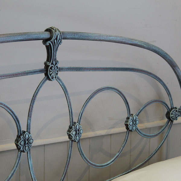 Close-up of a metal bed frame headboard with a distressed blue-gray finish and ornate scrollwork details, shown against a light wall.