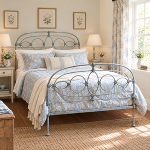 A styled bedroom scene featuring a metal bed frame with a blue and white floral patterned bedding set, cream throw blanket, matching curtains, and framed botanical prints on the wall.
