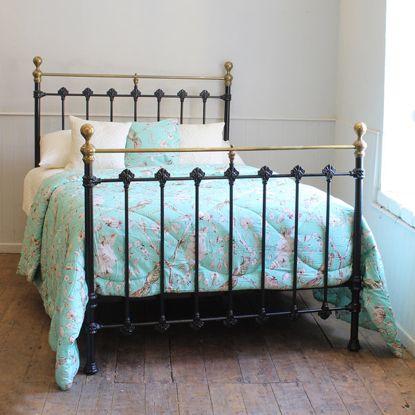 A vintage-style metal bed frame with a black wrought-iron style footboard and headboard accented by brass rails and gold-toned finials, dressed with a light blue floral quilt and white pillows in a bedroom setting.