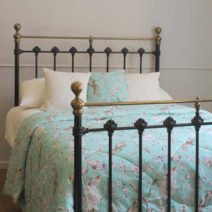 A bed with a black metal frame and brass accents, styled with a mint green floral comforter and matching pillows against a neutral wall.
