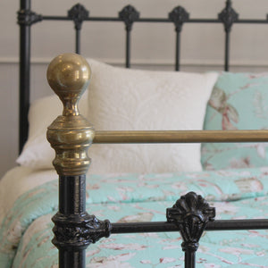 Close-up of a bed frame with a polished brass finial and decorative black metal rails, shown with white and mint floral bedding against a neutral wall.