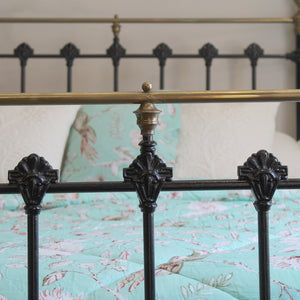 Close-up of a black metal bed frame with ornate scrollwork posts and a brass headboard rail, shown with a mint green floral-patterned quilt and white pillows in a bedroom setting.