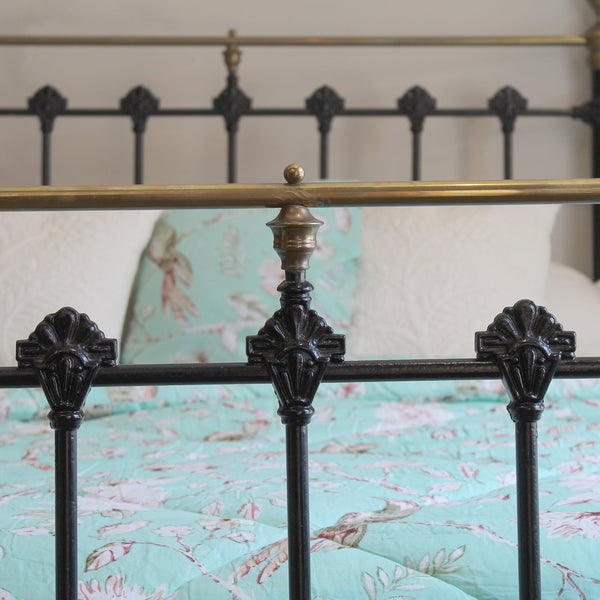 Close-up of a black metal bed frame with ornate scrollwork posts and a brass headboard rail, shown with a mint green floral-patterned quilt and white pillows in a bedroom setting.