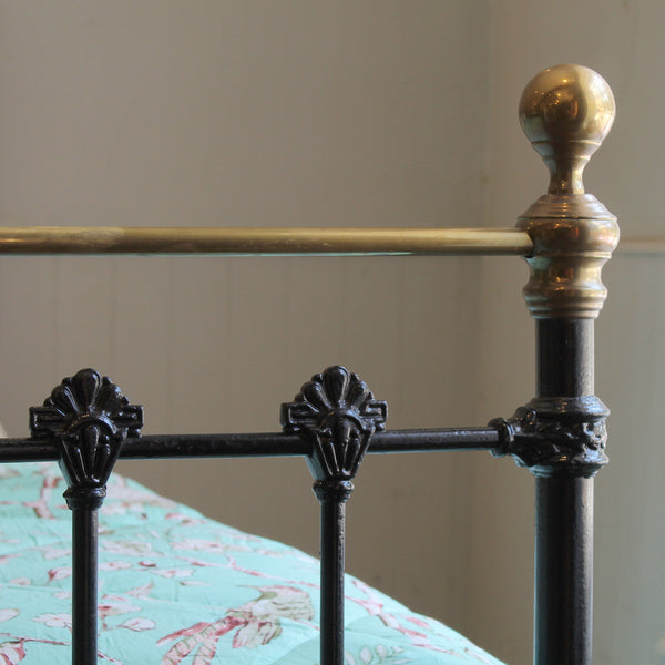 Close-up of an ornate metal bed frame with a black wrought-iron style body and a brass/gold-toned headboard rail and post cap, with part of a light blue floral quilt visible.