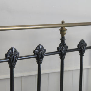 Close-up of a black metal bed frame rail with ornate decorative finials and a brass-colored horizontal support rod against a white paneled wall.