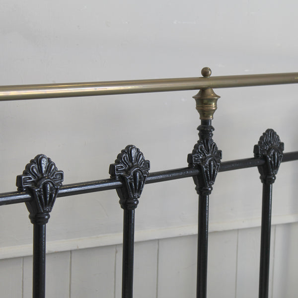 Close-up of a black metal bed frame rail with ornate decorative finials and a brass-colored horizontal support rod against a white paneled wall.