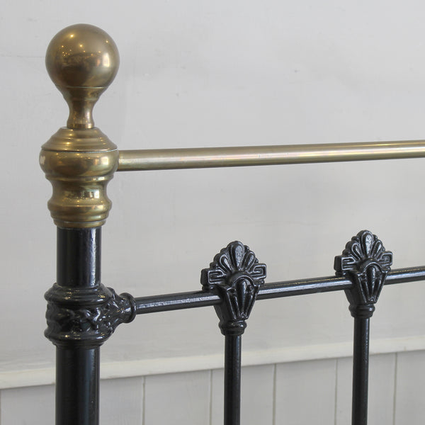 Close-up of a bed frame showing a polished brass finial on a black metal post, with horizontal rails and decorative scrollwork details against a plain light wall.