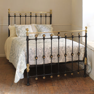 A black metal bed frame with ornate gold-toned accents and brass-finished posts, shown on a wooden floor in a bedroom setting with patterned bedding.